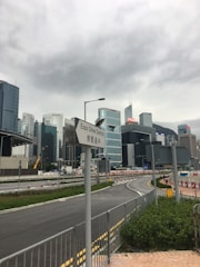 A cityscape featuring several modern skyscrapers and office buildings under a cloudy sky. A road with traffic signs, including one marked 'Expo Drive Central', is seen in the foreground. The area is surrounded by construction barriers and urban infrastructure.