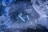 Two blue wireless earbuds are placed on a textured icy surface surrounded by pieces of ice, creating a cool and refreshing atmosphere.