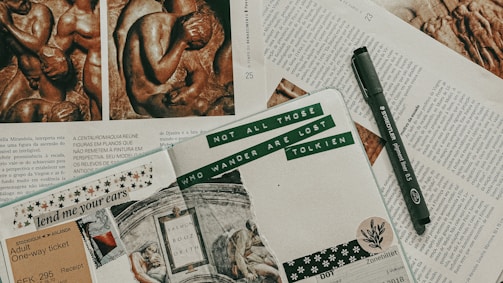 An artistic journal spread on top of several open magazine pages. The journal includes text, decorative stickers, a ticket stub, and a quote by Tolkien. Beside the journal is a black fine liner pen. The magazines feature images of sculptures and columns of text in a language that appears to be Portuguese.