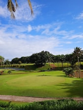A lush green golf course featuring a putting green with a flagstick in the center. In the background, there is a landscape of rolling hills adorned with palm trees and other vegetation. The sky above is bright blue with a few scattered clouds.