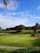 A lush green golf course featuring a putting green with a flagstick in the center. In the background, there is a landscape of rolling hills adorned with palm trees and other vegetation. The sky above is bright blue with a few scattered clouds.