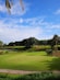 A lush green golf course featuring a putting green with a flagstick in the center. In the background, there is a landscape of rolling hills adorned with palm trees and other vegetation. The sky above is bright blue with a few scattered clouds.