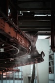A complex metal structure with interconnected pipes and beams, part of an industrial or mechanical setup, is visible. Steam or smoke is emanating from the equipment, which is colored dark brown and black.