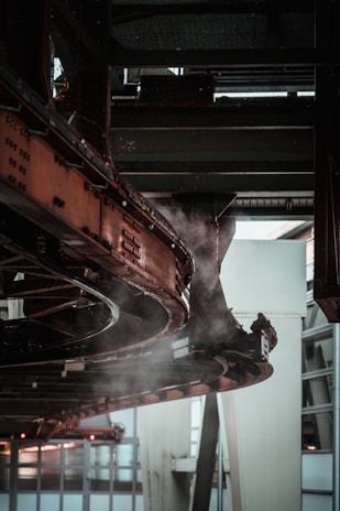 A complex metal structure with interconnected pipes and beams, part of an industrial or mechanical setup, is visible. Steam or smoke is emanating from the equipment, which is colored dark brown and black.