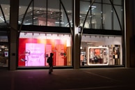 The image depicts a shop window display at night, featuring bright and colorful signage with the text 'FOR EVERY WISH THERE'S A GIFT' against a pink and red background. The interior of the store is visible through large windows, showing well-lit sections with various products. A silhouette of a person carrying a bag is walking past the store.
