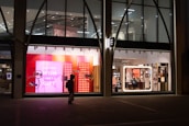 The image depicts a shop window display at night, featuring bright and colorful signage with the text 'FOR EVERY WISH THERE'S A GIFT' against a pink and red background. The interior of the store is visible through large windows, showing well-lit sections with various products. A silhouette of a person carrying a bag is walking past the store.