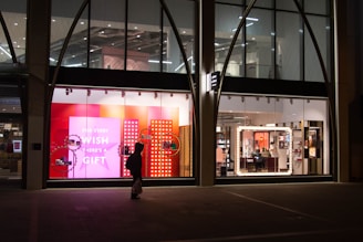 The image depicts a shop window display at night, featuring bright and colorful signage with the text 'FOR EVERY WISH THERE'S A GIFT' against a pink and red background. The interior of the store is visible through large windows, showing well-lit sections with various products. A silhouette of a person carrying a bag is walking past the store.