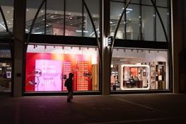 The image depicts a shop window display at night, featuring bright and colorful signage with the text 'FOR EVERY WISH THERE'S A GIFT' against a pink and red background. The interior of the store is visible through large windows, showing well-lit sections with various products. A silhouette of a person carrying a bag is walking past the store.