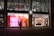The image depicts a shop window display at night, featuring bright and colorful signage with the text 'FOR EVERY WISH THERE'S A GIFT' against a pink and red background. The interior of the store is visible through large windows, showing well-lit sections with various products. A silhouette of a person carrying a bag is walking past the store.