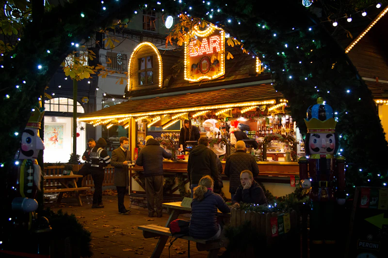 A lit-up wooden holiday market bar stall strung with lights, with visitors gathered at picnic tables (generic Christmas market photo, not verifiably Chicago's Christkindlmarket)