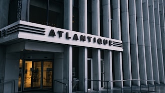 The entrance to a building with the name 'Atlantique' prominently displayed above glass doors. The architectural design features vertical lines and metallic accents, giving it a modern and sleek appearance. Subdued lighting comes from inside, contrasting with the exterior's gray tones.