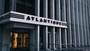 The entrance to a building with the name 'Atlantique' prominently displayed above glass doors. The architectural design features vertical lines and metallic accents, giving it a modern and sleek appearance. Subdued lighting comes from inside, contrasting with the exterior's gray tones.