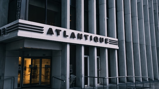 The entrance to a building with the name 'Atlantique' prominently displayed above glass doors. The architectural design features vertical lines and metallic accents, giving it a modern and sleek appearance. Subdued lighting comes from inside, contrasting with the exterior's gray tones.