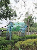 Two blue abstract sculptures with intricate designs are set amidst lush greenery, near a building with a geometric green facade. The scene is surrounded by vibrant trees and shrubs, creating a harmonious blend of art and nature.