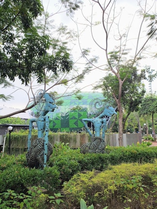 Two blue abstract sculptures with intricate designs are set amidst lush greenery, near a building with a geometric green facade. The scene is surrounded by vibrant trees and shrubs, creating a harmonious blend of art and nature.