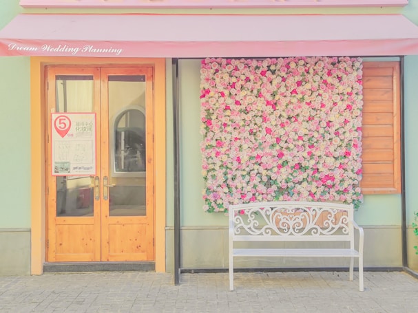 A charming storefront featuring a decorative wall covered in pink and white roses alongside a wooden door with glass panels. A white ornamental bench sits on the right, under a soft pink awning with 'Dream Wedding Planning' written in cursive.
