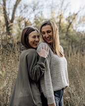Two women are standing close together in an outdoor setting, surrounded by tall, dried plants and trees with autumn foliage in the background. Both are smiling and seem to be enjoying each other's company. They are wearing casual sweaters, radiating a sense of friendship and happiness.