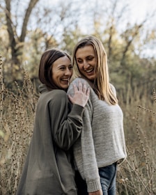 Two women are standing close together in an outdoor setting, surrounded by tall, dried plants and trees with autumn foliage in the background. Both are smiling and seem to be enjoying each other's company. They are wearing casual sweaters, radiating a sense of friendship and happiness.