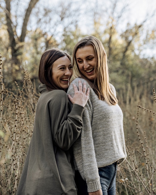 Two women are standing close together in an outdoor setting, surrounded by tall, dried plants and trees with autumn foliage in the background. Both are smiling and seem to be enjoying each other's company. They are wearing casual sweaters, radiating a sense of friendship and happiness.