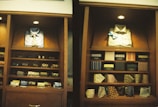 A display of clothing and accessories in a wooden cabinet, featuring neatly arranged shirts, ties, and boxes. The left side showcases a blue button-up shirt with a patterned tie, while the right side displays a light-colored shirt with a blue bow tie. Below are shelves with various patterned and colored ties, and some boxed items.