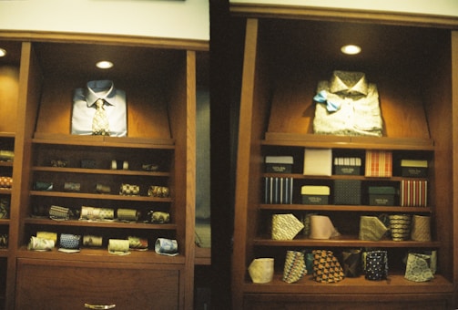 A display of clothing and accessories in a wooden cabinet, featuring neatly arranged shirts, ties, and boxes. The left side showcases a blue button-up shirt with a patterned tie, while the right side displays a light-colored shirt with a blue bow tie. Below are shelves with various patterned and colored ties, and some boxed items.