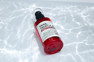 A red-capped bottle labeled 'Snail Truecica Miracle Repair Serum' lies on a surface with a pattern resembling light reflections on water. The bottle is clear with red accents, containing a dark liquid inside.