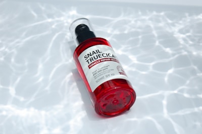 A red-capped bottle labeled 'Snail Truecica Miracle Repair Serum' lies on a surface with a pattern resembling light reflections on water. The bottle is clear with red accents, containing a dark liquid inside.