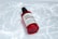 A red-capped bottle labeled 'Snail Truecica Miracle Repair Serum' lies on a surface with a pattern resembling light reflections on water. The bottle is clear with red accents, containing a dark liquid inside.