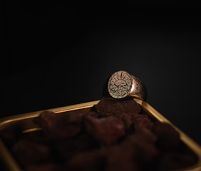 A gold ring with an engraved design of mountains and a setting sun is resting on a pile of small stones. The scene is set against a dark background, highlighting the intricate details of the ring.