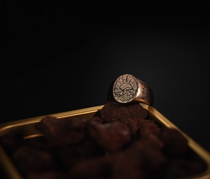 A gold ring with an engraved design of mountains and a setting sun is resting on a pile of small stones. The scene is set against a dark background, highlighting the intricate details of the ring.