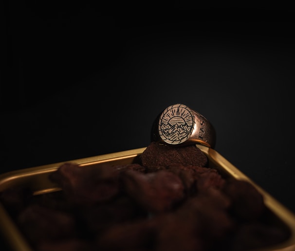 A gold ring with an engraved design of mountains and a setting sun is resting on a pile of small stones. The scene is set against a dark background, highlighting the intricate details of the ring.
