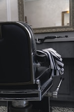 A classic black leather barber chair with shiny metal details is positioned in front of a countertop holding grooming tools. A striped barber cape is draped over the chair's armrest. A large, ornate mirror hangs above the countertop in a room with subdued lighting, reflecting part of the room.