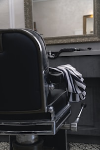 A classic black leather barber chair with shiny metal details is positioned in front of a countertop holding grooming tools. A striped barber cape is draped over the chair's armrest. A large, ornate mirror hangs above the countertop in a room with subdued lighting, reflecting part of the room.