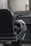 A classic black leather barber chair with shiny metal details is positioned in front of a countertop holding grooming tools. A striped barber cape is draped over the chair's armrest. A large, ornate mirror hangs above the countertop in a room with subdued lighting, reflecting part of the room.