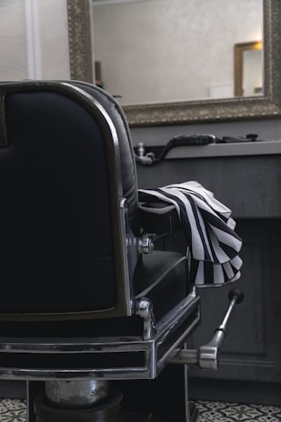 A classic black leather barber chair with shiny metal details is positioned in front of a countertop holding grooming tools. A striped barber cape is draped over the chair's armrest. A large, ornate mirror hangs above the countertop in a room with subdued lighting, reflecting part of the room.