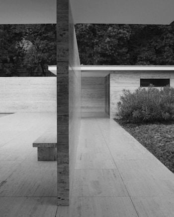 A modern architectural structure with clean lines and a minimalist design. The building features large travertine walls and floors, with a central dividing wall and a built-in bench. There's a garden area with lush green plants to the right, and dense trees in the background. The overall setting appears serene and elegant.