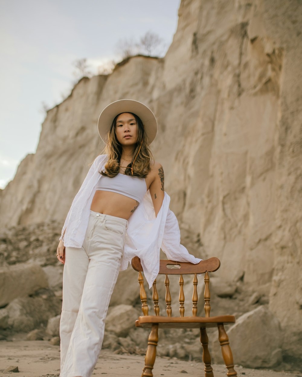 a woman in a white top and white pants standing next to a wooden chair