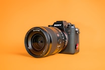 A professional Sony camera with a large lens is positioned on an orange background. The camera, a Sony Alpha model, features detailed branding and controls visible on its body.
