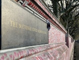 A memorial wall stretches into the distance, covered with numerous red hearts. A dark plaque with gold lettering reads, 'The National Covid Memorial Wall.' The wall is adorned with messages and pictures, and there are trees lining the path alongside it.
