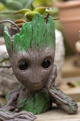 A detailed figure of a small tree-like character with large expressive eyes, and green foliage sprouting from the top of its head. The figure appears to be sitting with one hand resting on its cheek, giving a thoughtful or curious expression. The background includes blurred elements of potted plants and foliage.