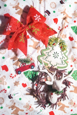 A collection of Christmas-themed decorations placed on a festive fabric featuring snowmen and reindeer patterns. The central items include a large red bow with snowflake and holly designs, a small decorative red car with a Christmas tree on top, a green and white Christmas tree ornament, and a handmade reindeer ornament within a rustic wreath. The background fabric enhances the holiday theme with illustrations like candy canes and Santa hats.
