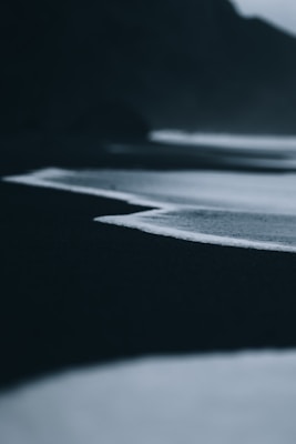 A dark, moody coastline with gentle waves creating a white outline against the black sand. The background is blurred, adding to the mysterious atmosphere.