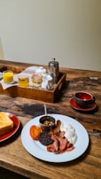 A hearty breakfast is arranged on a rustic wooden table. On a white plate are fried eggs, bacon, sausage, black pudding, grilled tomato, and baked beans in a small cup. Nearby, slices of toast are served on a red plate. A wooden tray holds glass jars, a metal coffee pot, and creamers. A red bowl and saucer are set beside the tray.