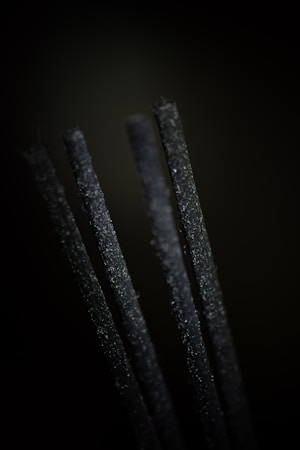 Four upright black incense sticks are in close-up focus against a dark background, emphasizing their textured surfaces.