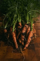 Several freshly harvested carrots with leafy tops are lying on a wooden surface, covered in soil. The scene suggests freshness and natural production.
