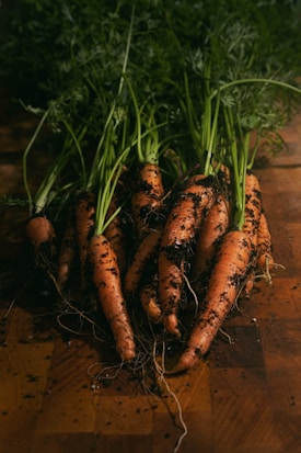 Several freshly harvested carrots with leafy tops are lying on a wooden surface, covered in soil. The scene suggests freshness and natural production.