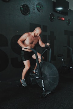 A man is intensely working out on a stationary exercise bike in a gym setting. He is shirtless and wearing black shorts and sneakers. The environment is dimly lit with dark walls adorned with workout equipment such as weights. A digital clock displays zero on the wall, and there is a television screen mounted above.