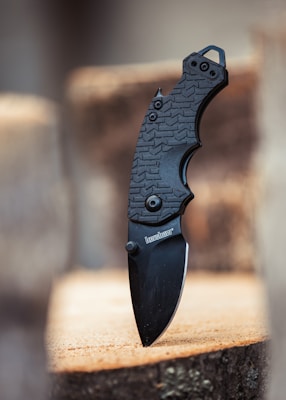 A black folding knife with a textured handle is embedded into a wooden surface. The knife has a sleek design and features a visible brand logo on the blade.