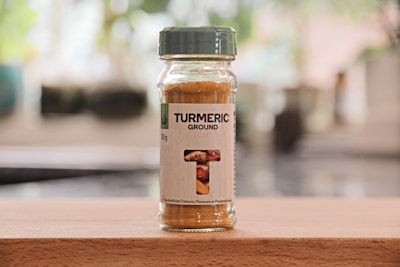 A glass jar of ground turmeric with a green lid is placed on a wooden surface. The label features the words 'Turmeric Ground' along with a small image of turmeric roots.