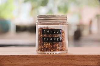 A glass jar with a silver lid contains reddish-brown chilli flakes. The jar is labeled with black tape displaying the words 'CHILLI FLAKES.' It is placed on a wooden surface, with a blurred, green and brown background suggesting a natural or kitchen setting.